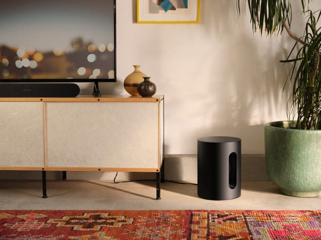 Sonos Sub mini zwart gepositioneerd naast een tv meubel in een kleurrijke woonkamer.